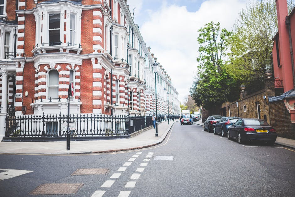 A quiet residential street in Islington with a row of Victorian-style terraced houses made of red and white brickwork. The buildings feature decorative architectural details including bay windows, ornate window trims, and black wrought iron railings along the pavement. Several parked cars, including a black sedan and a silver vehicle, are lined up along the right side of the street. The pavement is clear, with a few bollards and road markings visible, such as dashed white lines indicating parking restrictions. In the background, a moving van is partially visible near the curb, parked in front of the houses, with the loading process possibly underway as part of home relocation or furniture transport activities. Bright natural daylight illuminates the scene, with green trees lining the sidewalks, and a slightly cloudy sky overhead. This image relates to house removals or moving services offered by Removal Van Islington, emphasizing urban moving logistics and packing considerations.