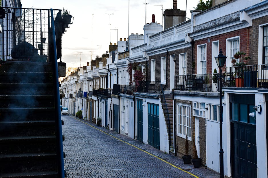 A quiet residential street in Islington with a row of Victorian-style terraced houses made of red and white brickwork. The buildings feature decorative architectural details including bay windows, ornate window trims, and black wrought iron railings along the pavement. Several parked cars, including a black sedan and a silver vehicle, are lined up along the right side of the street. The pavement is clear, with a few bollards and road markings visible, such as dashed white lines indicating parking restrictions. In the background, a moving van is partially visible near the curb, parked in front of the houses, with the loading process possibly underway as part of home relocation or furniture transport activities. Bright natural daylight illuminates the scene, with green trees lining the sidewalks, and a slightly cloudy sky overhead. This image relates to house removals or moving services offered by Removal Van Islington, emphasizing urban moving logistics and packing considerations.