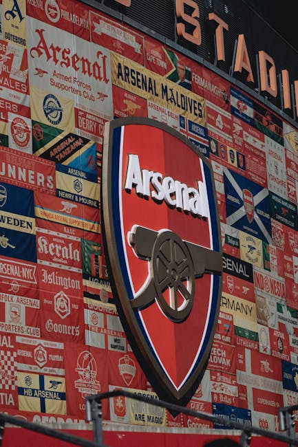 A large, illuminated Arsenal Football Club crest is mounted on a wall covered with a collage of various vintage football banners, logos, and memorabilia. The crest features a red shield with a white border, the word 'Arsenal' in bold white letters at the top, and a black cannon facing to the right. The wall behind is densely decorated with colorful fabric and paper banners, including team names, slogans, and sponsor logos, arranged in overlapping layers. The scene appears to be outdoors, possibly in the stadium or a memorabilia display area, with part of a metal railing visible at the bottom of the image. This setting reflects a passion for football and club history, aligning with sports-related themes often associated with football clubs like Arsenal, though it is not directly related to house removals or moving services.