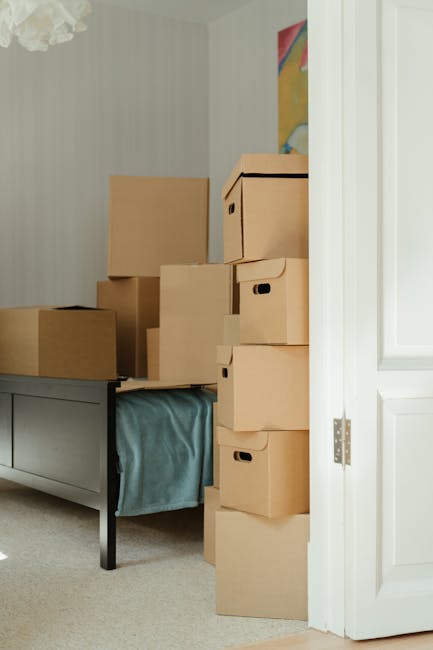 A stack of brown cardboard boxes of various sizes are arranged on and around a grey piece of furniture inside a room, with some boxes placed on top of each other and others leaning against the furniture. Several of the boxes have carry handles cut out, and they are packed with packing materials. The room features light-colored walls with a subtle striped pattern, and a small section of a colorful abstract painting is visible on the wall. Part of a white door frame and a partially open door are visible on the right side of the image. The packed boxes are prepared to be moved as part of a home relocation process, and the scene is illuminated by natural light, reflecting the loading or packing phase in the moving process. The environment and furniture arrangement suggest that Removal Van Islington provides professional removals, including packing, loading, and transporting household items.