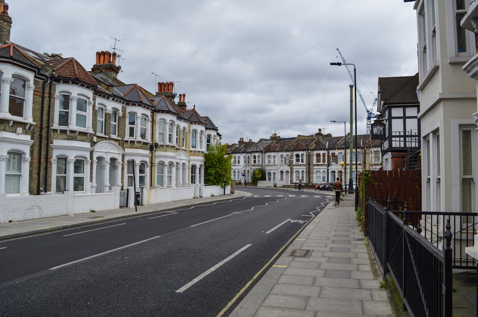 Wide street scene on Essex Road in Islington, featuring Victorian-style terraced houses with white and brick facades, bay windows, and red-tiled roofs. The pavement runs along the right side next to residential properties with black metal railings and some greenery. A tall street lamp and a construction crane are visible against an overcast sky, indicating urban activity. The road has marked parking bays and is currently empty of vehicles. This image relates to house removals and relocation services by Removal Van Islington, suggesting a typical residential area where furniture transport and packing processes might be taking place during a home relocation or moving operation.