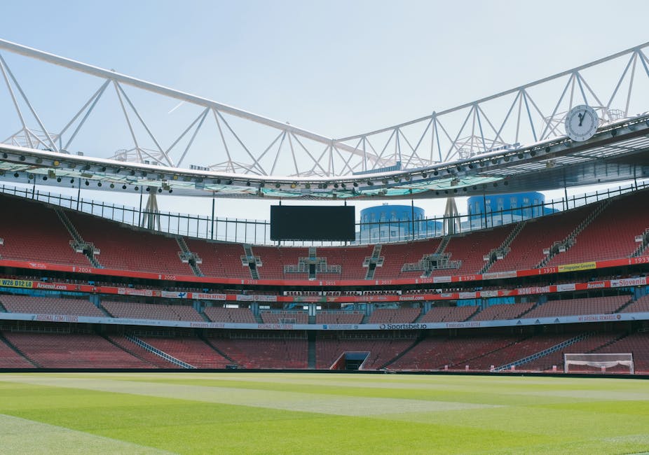Interior view of a large sports stadium with a partially open roof structure supported by white steel trusses, allowing natural daylight to illuminate the scene. The field is visible with neatly maintained green grass, surrounded by empty red seating sections arranged in tiers. Above the seating, multiple rows of digital advertising boards display various messages and logos, including some references to football teams and sponsors. A tall, centrally mounted digital scoreboard is situated above the seating, currently turned off. The perimeter of the stadium includes access walkways, railings, and structural elements. In the background, external building structures and a blue sky with minimal cloud cover are visible, indicating a clear day. This setting relates to event spaces such as sports venues, and the image could depict the environment involved in large-scale events requiring logistics, including equipment or furniture transport for event setup or house removals related to stadium staff or equipment.