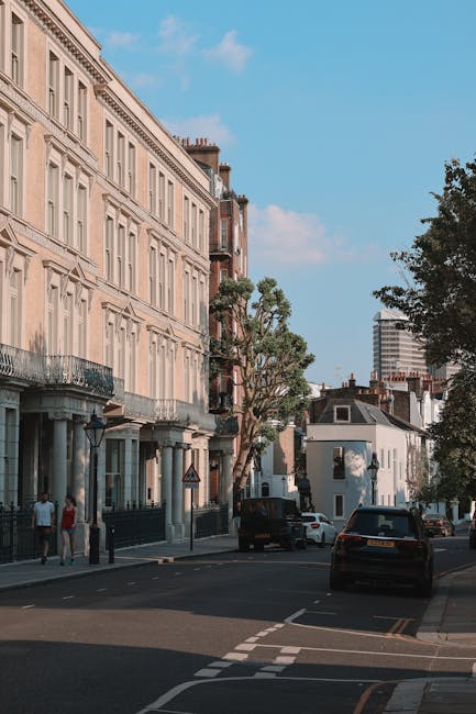 A street scene in an urban residential area with multi-story white and beige terrace houses featuring large windows, decorative railings, and ornate moldings. In the foreground, a black van from Removal Van Islington is parked on the curb, with a few cardboard boxes and plastic-wrapped furniture pieces being loaded into it by movers not visible in the image. The sidewalk is occupied by a few pedestrians. Tall trees with leafy branches line the pavement, providing shade along the street, with a partly cloudy blue sky overhead. In the background, there are modern buildings and a few parked cars, indicating a busy yet organized neighbourhood typical of London. The overall scene reflects elements of home relocation and furniture transport associated with professional removals services in Islington, near Angel Upper Street.