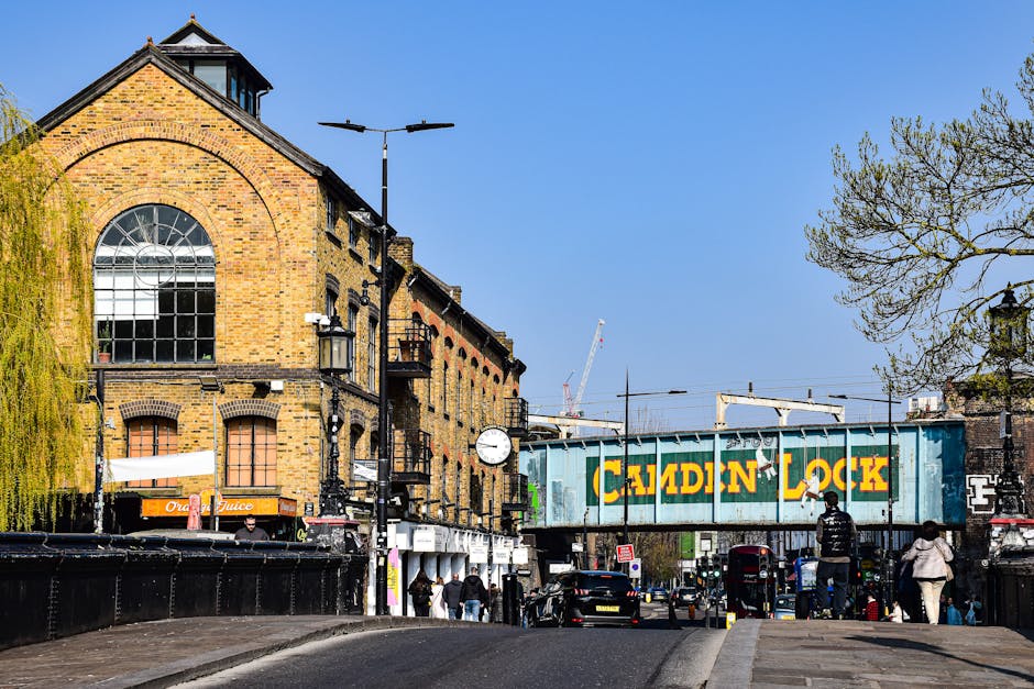 A daytime street scene in Islington near Angel Upper Street featuring a brick building with large arched windows, a clock mounted on a lamppost, and a Camden Lock railway freight container suspended on a bridge above the road. The street is busy with pedestrians walking along the pavement, some carrying shopping bags, and vehicles parked or moving along the road. Trees with budding leaves are visible on the right side, and in the background, construction cranes and overhead tram lines can be seen. The scene depicts an active urban environment typical of a home relocation or moving logistics context, with [COMPANY_NAME] providing removals services in the area, as part of packing, loading, and furniture transport processes.