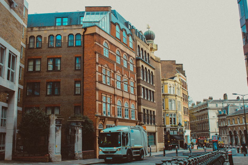 A street scene in an urban residential area with multi-story white and beige terrace houses featuring large windows, decorative railings, and ornate moldings. In the foreground, a black van from Removal Van Islington is parked on the curb, with a few cardboard boxes and plastic-wrapped furniture pieces being loaded into it by movers not visible in the image. The sidewalk is occupied by a few pedestrians. Tall trees with leafy branches line the pavement, providing shade along the street, with a partly cloudy blue sky overhead. In the background, there are modern buildings and a few parked cars, indicating a busy yet organized neighbourhood typical of London. The overall scene reflects elements of home relocation and furniture transport associated with professional removals services in Islington, near Angel Upper Street.