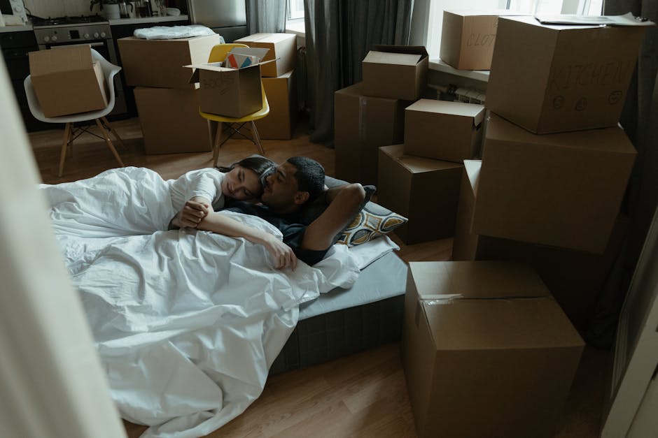A couple lying together on a bed inside a room filled with numerous cardboard moving boxes of varying sizes, some sealed with packing tape and others open. The woman is resting her head on the man's s
