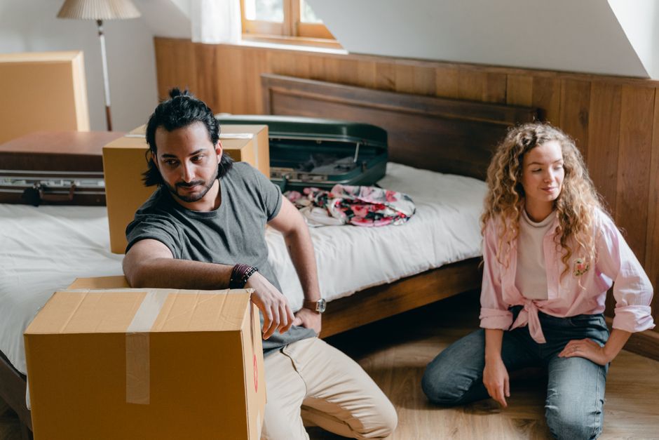 A couple lying together on a bed inside a room filled with numerous cardboard moving boxes of varying sizes, some sealed with packing tape and others open. The woman is resting her head on the man's s