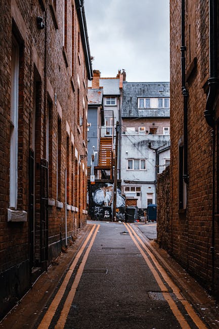 A narrow alleyway between two brick buildings in Islington, with the pavement marked by double yellow lines. The alley leads to a small, cluttered back area of a residential property, featuring a wooden staircase attached to a gray concrete building, with windows, rooftop structures, and rooftop chimneys visible. The ground is wet, reflecting the overcast sky above. In the background, various items associated with home relocation and furniture transport, such as plastic-covered furniture, cardboard boxes, and packing materials, are stacked near the rear entrance. Equipment like trolleys, straps, or blankets may be used in the loading process, as a professional removals service like Removal Van Islington navigates tight access points for house removals or moving logistics.