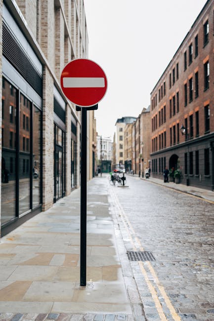 The image depicts a narrow urban street with a red and white 'no entry' traffic sign mounted on a black pole positioned on a stone-paved sidewalk. To the left, there are multi-storey buildings with large glass windows and brick facades, suggesting a commercial or residential area. On the right, more brick buildings line the street, which appears to be a one-way street based on the sign and the absence of vehicles facing the opposite direction. In the background, there are a few pedestrians walking along the pavement, and some cyclists are visible further down the street. The street surface is cobblestone, with double yellow lines along the curb indicating parking restrictions. The scene is well-lit with natural daylight, and the overall setting reflects a typical city environment suitable for home relocation or moving services, such as those provided by Removal Van Islington, operating in the area. Visibility of the street layout, building facades, and pedestrians supports planning for navigating narrow streets during furniture transport or packing and moving activities.