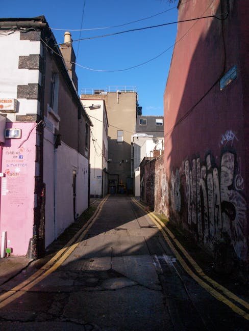 A narrow alleyway between two brick buildings in Islington, with the pavement marked by double yellow lines. The alley leads to a small, cluttered back area of a residential property, featuring a wooden staircase attached to a gray concrete building, with windows, rooftop structures, and rooftop chimneys visible. The ground is wet, reflecting the overcast sky above. In the background, various items associated with home relocation and furniture transport, such as plastic-covered furniture, cardboard boxes, and packing materials, are stacked near the rear entrance. Equipment like trolleys, straps, or blankets may be used in the loading process, as a professional removals service like Removal Van Islington navigates tight access points for house removals or moving logistics.
