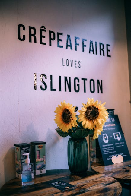 A close-up interior view of a table inside a premises shows a dark green glass vase containing three large, vibrant yellow sunflowers with dark centers. Next to the vase, there are bottles of hand sanitizer and promotional leaflets, some of which include illustrations of cleaning products. The wall behind features a sign with the words 'CRÊPE AFFAIRE LOVES ISLINGTON' in black, blue, and purple lettering, with the phrase 'LOVES' placed between the two larger words. The scene is lit with warm, natural lighting, highlighting the textured wooden surface of the table and the bright colors of the flowers. This setting appears to be part of a promotional or decorative display, possibly within a café or dedicated event space involved in a house or business relocation process, aligned with [COMPANY_NAME]'s removals and moving services in Islington.